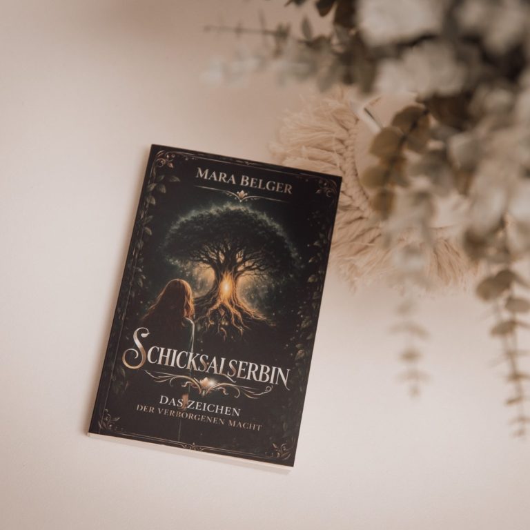 Ein Buch mit dem Titel „Schicksalserbin“ steht auf einem Tisch, umgeben von Pflanzen und Arbeitsmaterial.
JonahLu-Fotografie